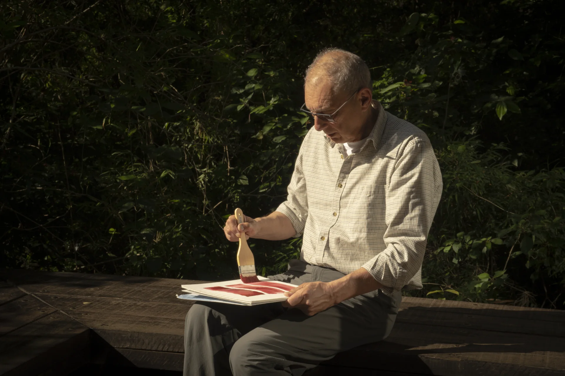 Older man painting with a brush on paper while sitting on a bench outdoors, surrounded by greenery.