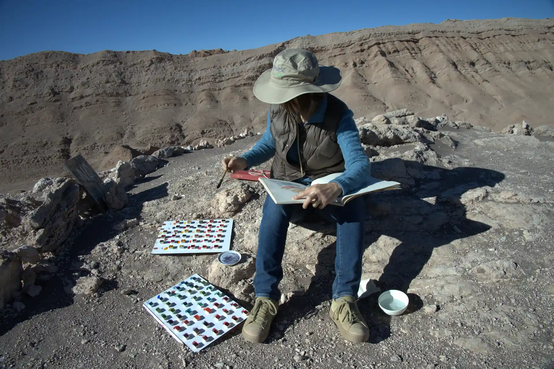 Person painting outdoors on rocky terrain, using a color palette and sketchbook under a clear blue desert sky.