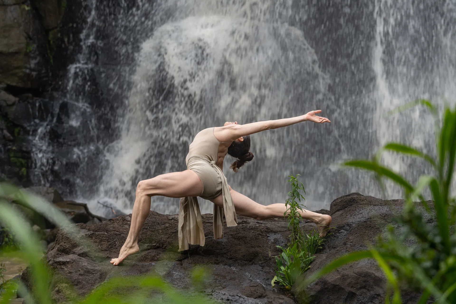 Dancer performs a graceful yoga pose on rocks beside a waterfall, surrounded by lush greenery.