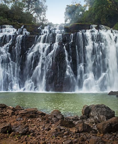 Wide waterfall cascading over dark rocks into a green pool, surrounded by trees and a rocky shoreline.