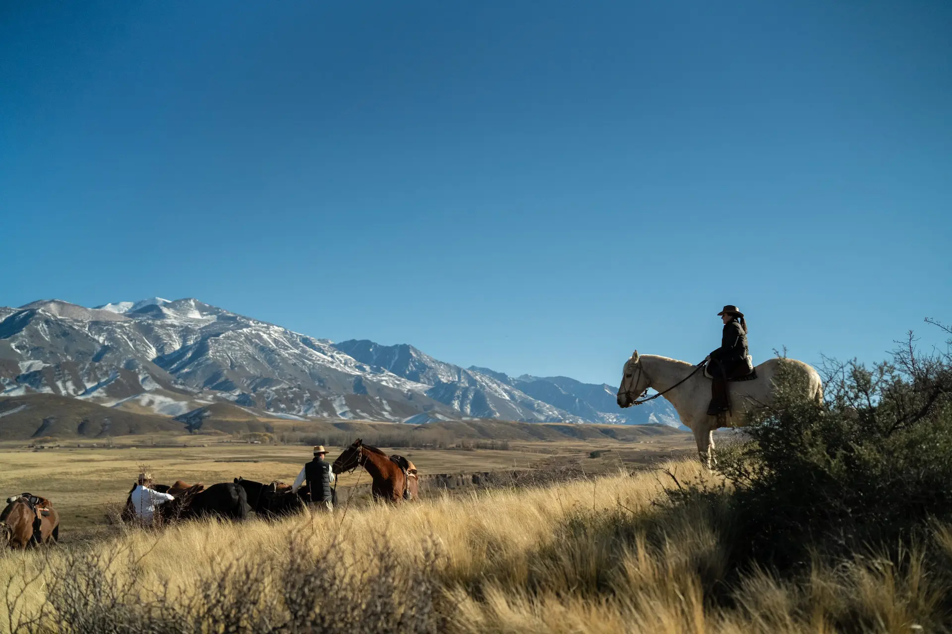 Riders on horseback crossing open grassland with snow-capped mountains in the distance under a clear blue sky.