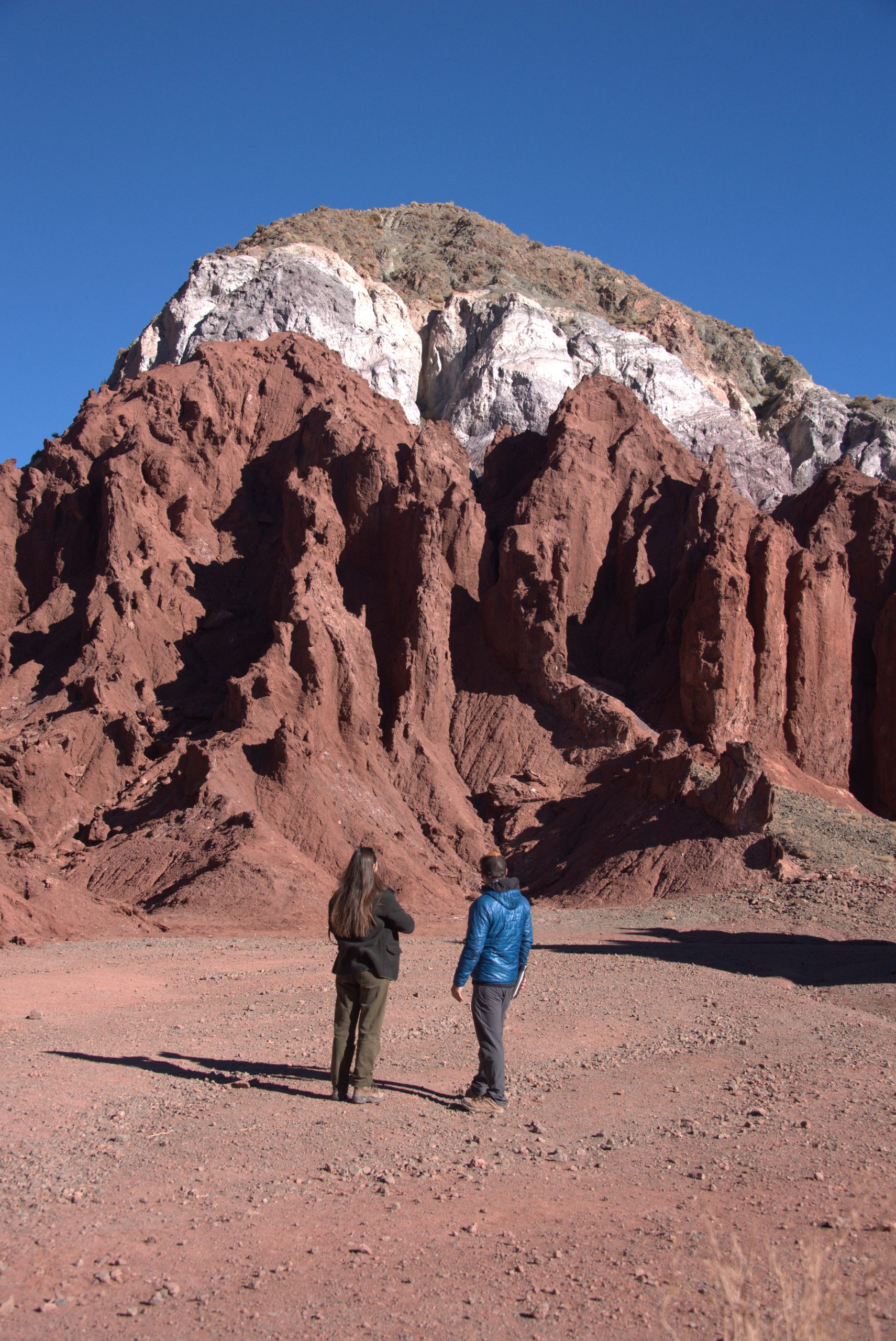 Two people standing on a desert plain, looking up at towering red and white rock formations.