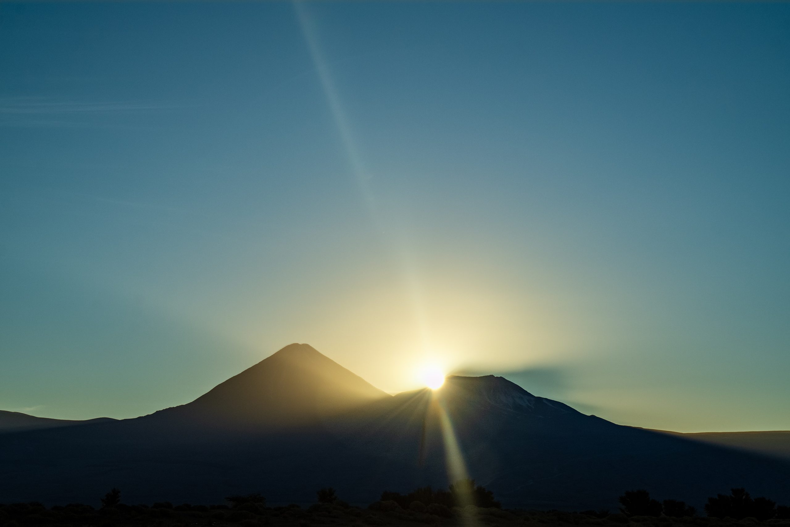 Sunrise behind a mountain ridge, casting soft rays and silhouettes against a clear blue sky.