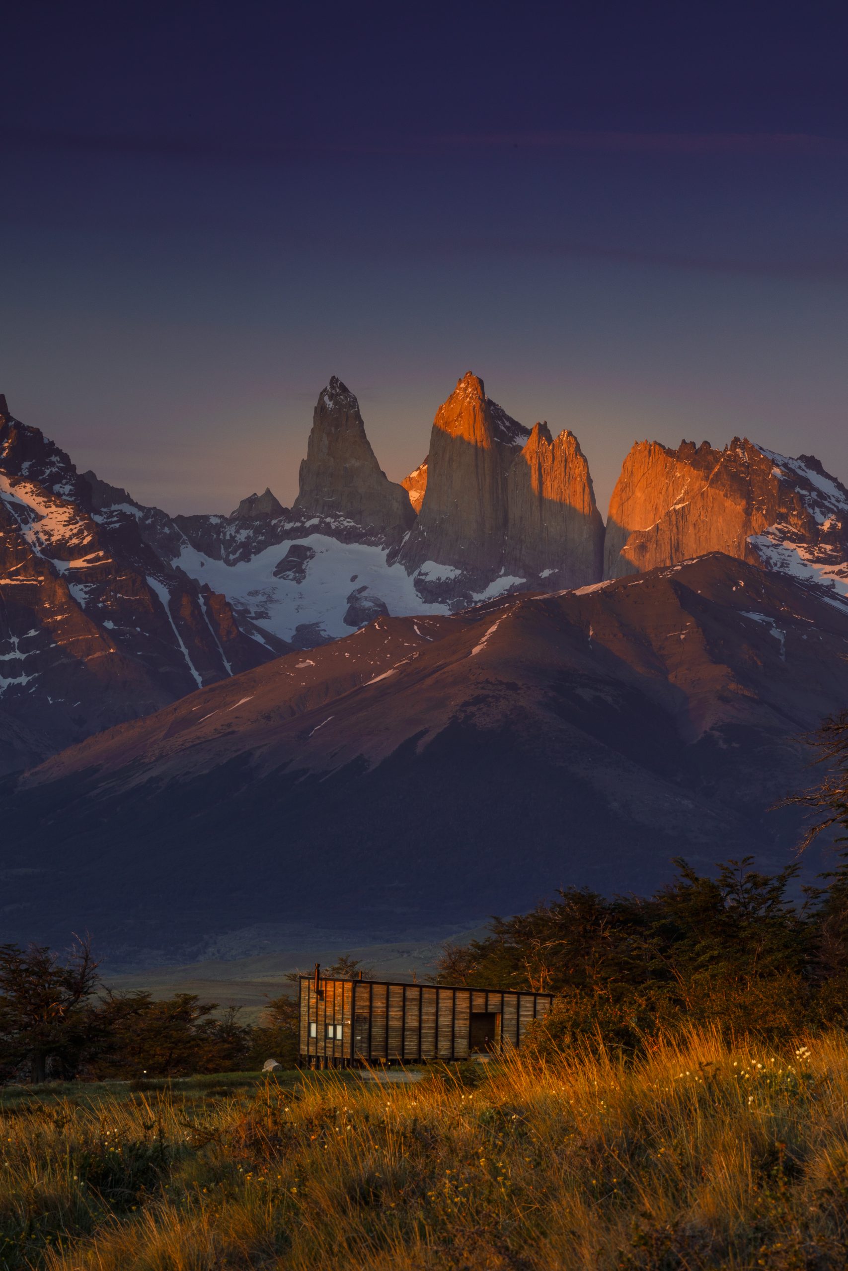 Sunlit mountain peaks rise behind a small cabin in a grassy valley at dusk under a deep blue sky.