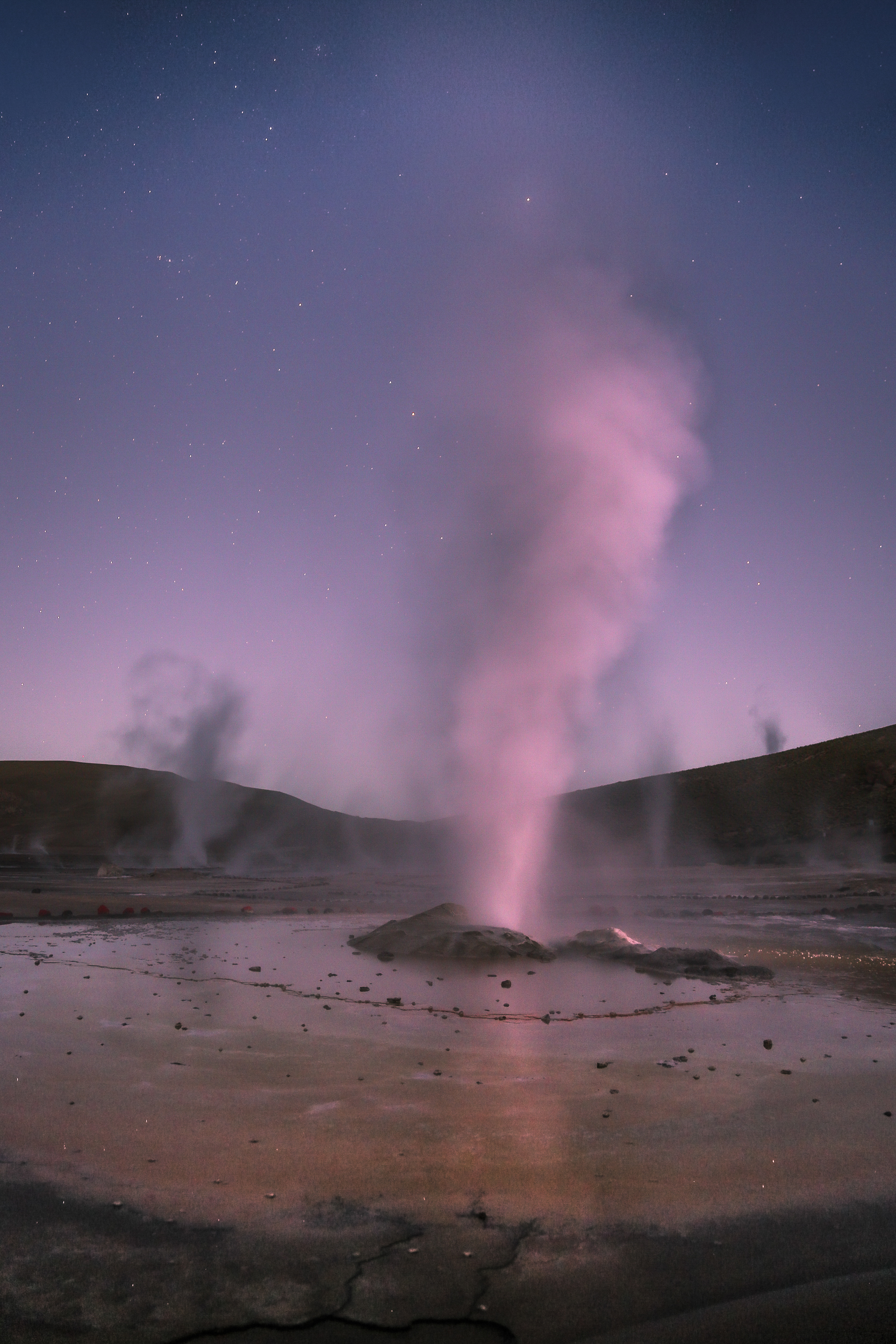 Steam vents rise from a geothermal field at dusk, glowing pink beneath a star-filled twilight sky.