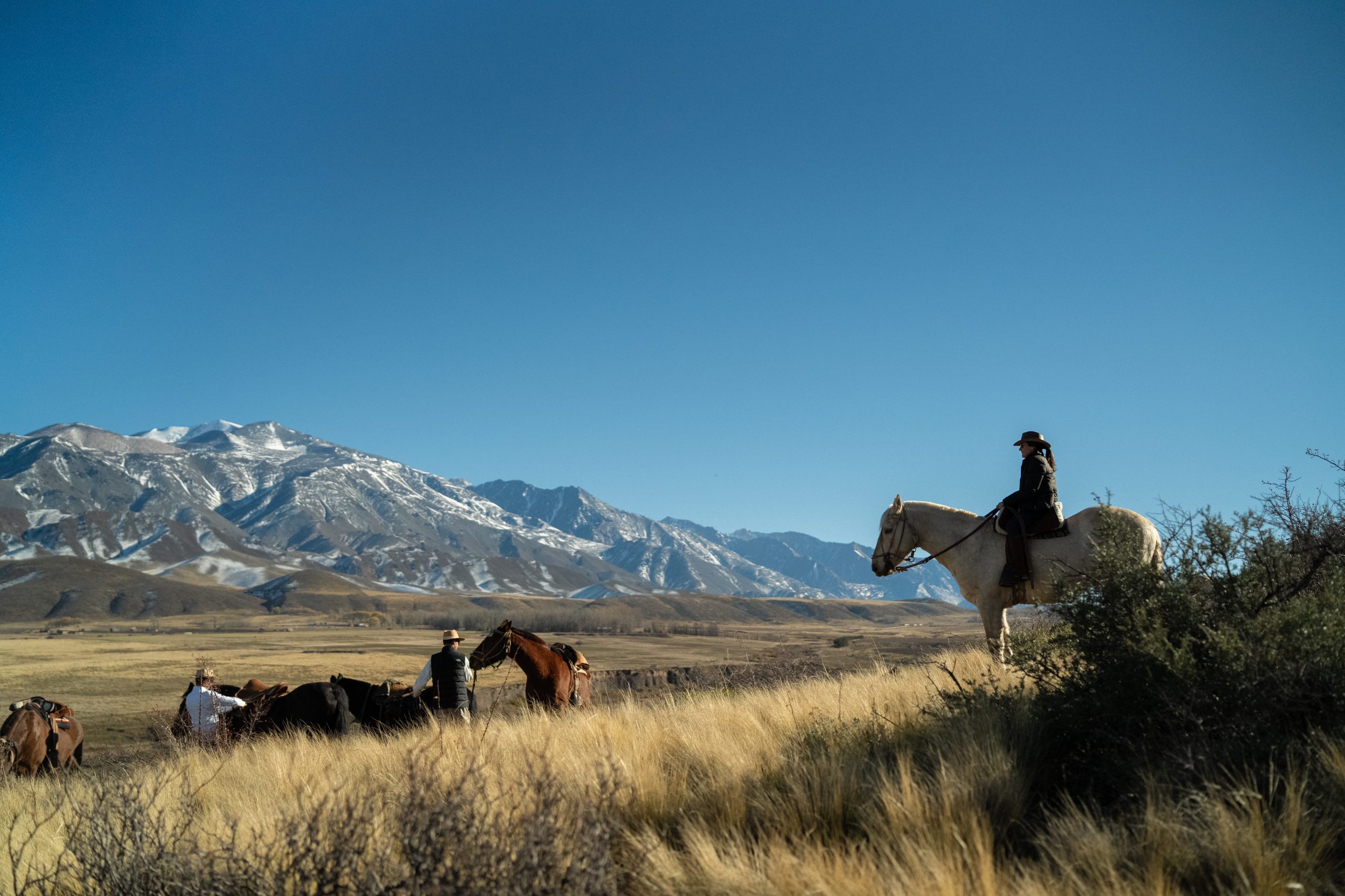Riders on horseback crossing open grassland with snow-capped mountains in the distance under a clear blue sky.