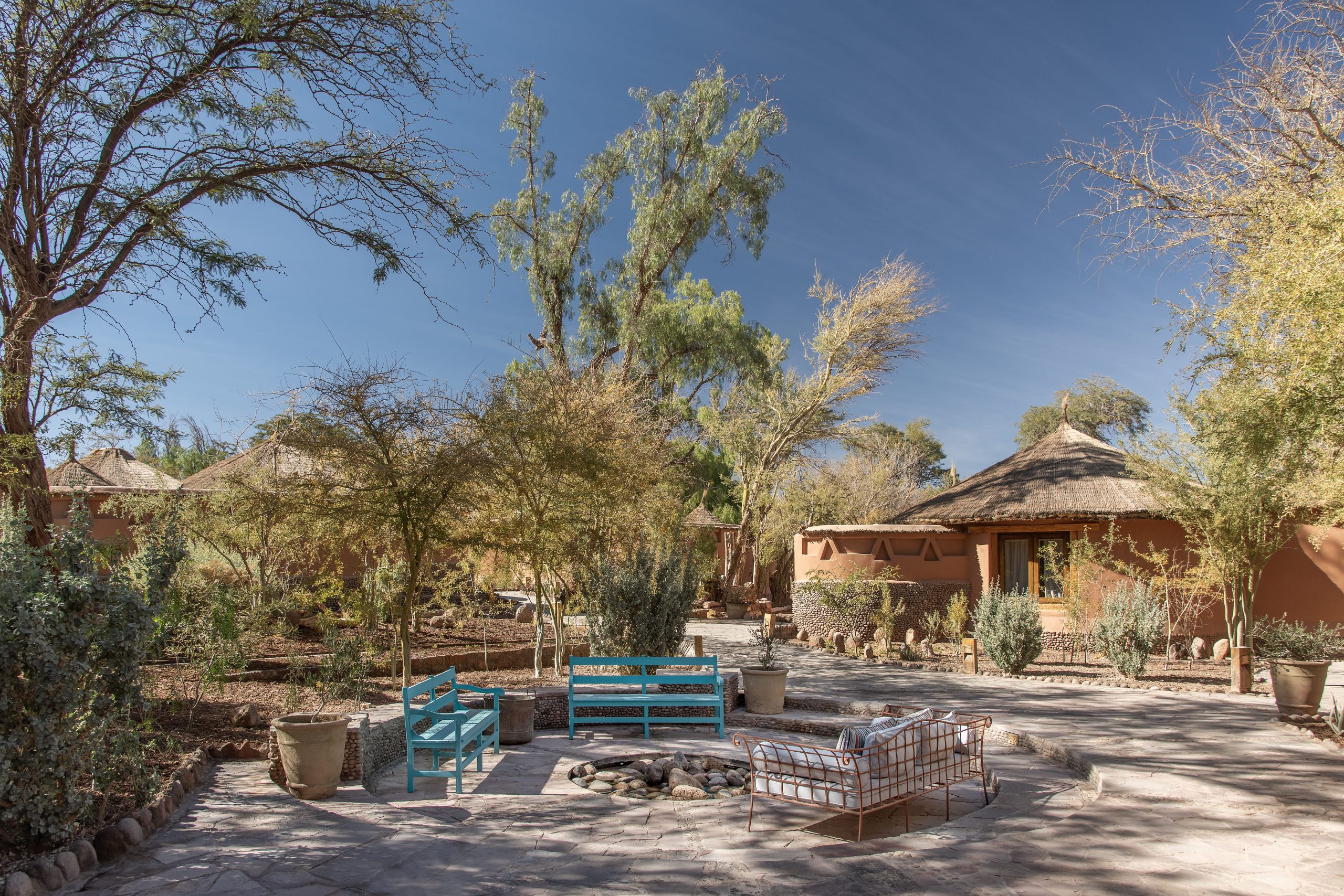 Desert lodge courtyard with adobe huts, thatched roofs, trees, and outdoor seating under a clear blue sky.