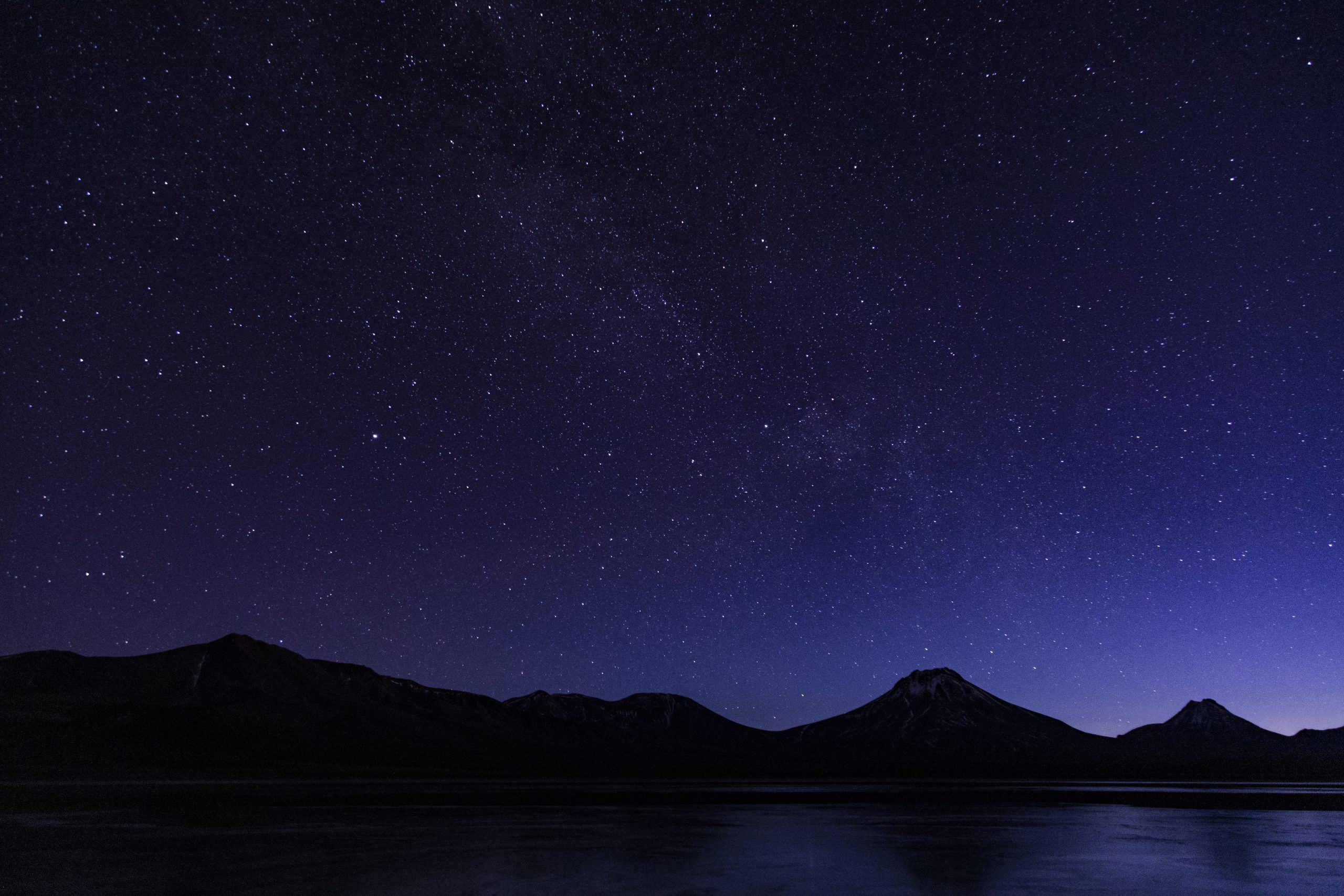 Star-filled night sky above dark mountain silhouettes reflected on calm water under a deep blue horizon.