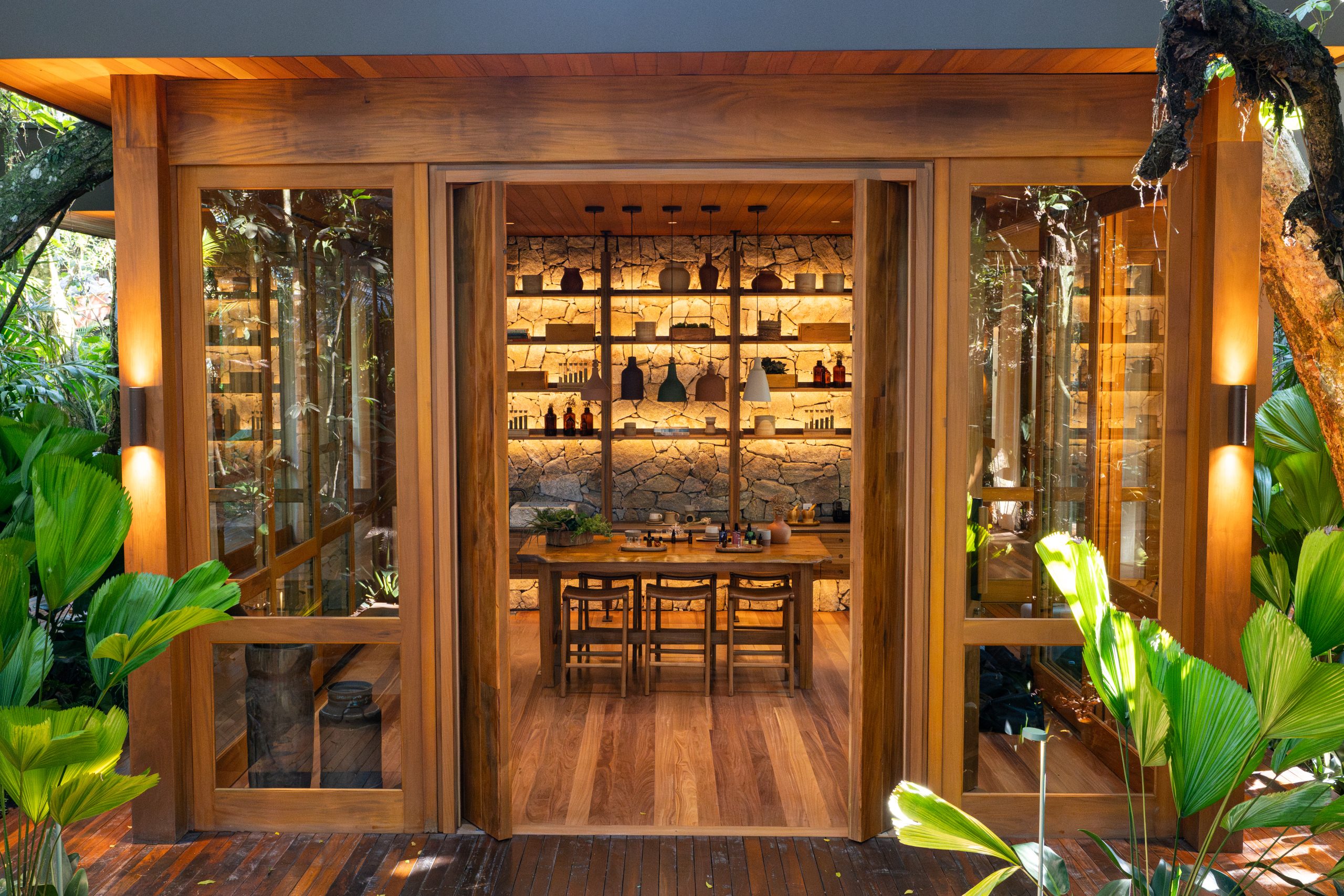 Warm wood pavilion with open glass doors revealing a dining table and shelves, surrounded by lush tropical greenery.
