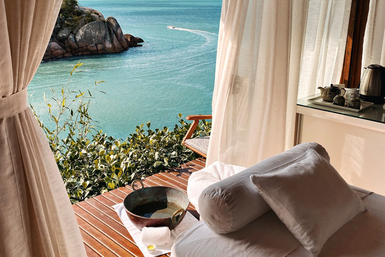 Ocean-view room with open curtains, wooden deck, soaking tub, and turquoise sea with rocky shoreline beyond.