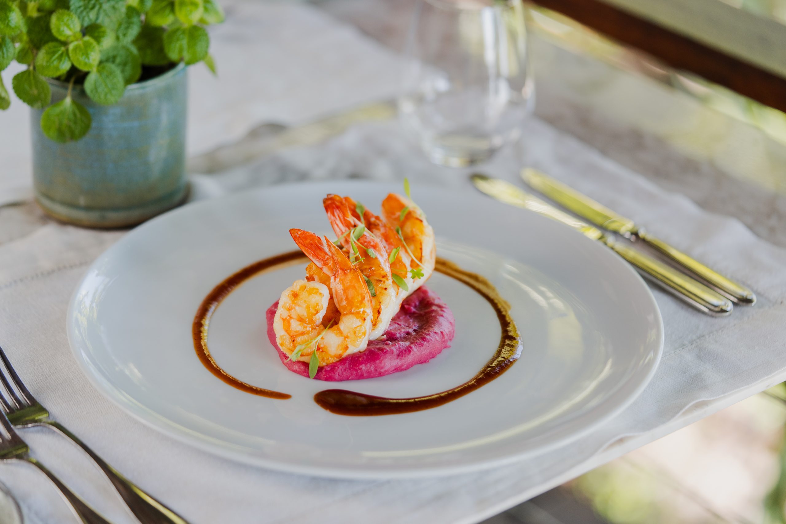 Plated shrimp served on pink purée with herb garnish and sauce, set on a white table with cutlery.