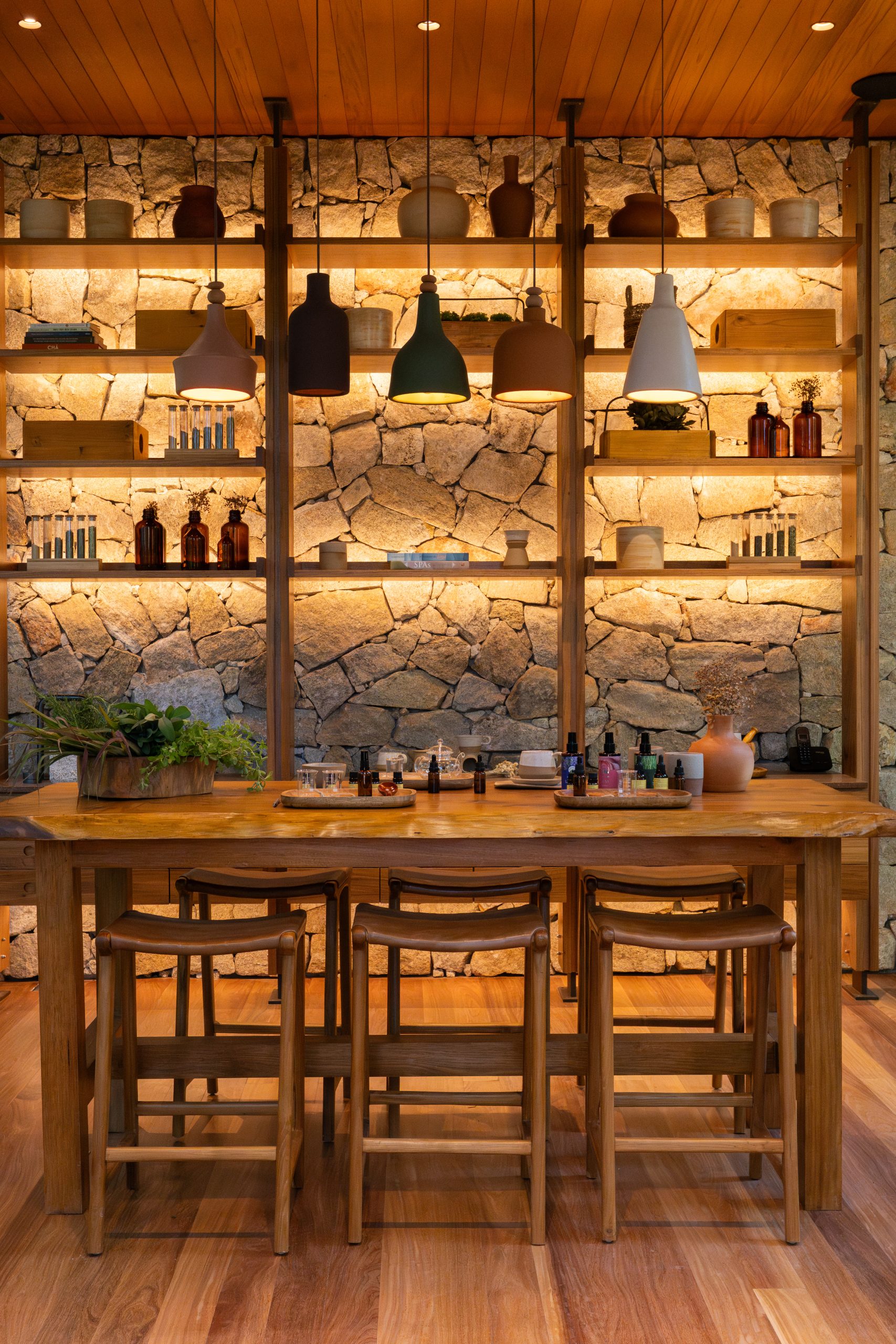 Warm wooden bar with stools, stone wall shelving, hanging pendant lights, and neatly arranged bottles and decor.