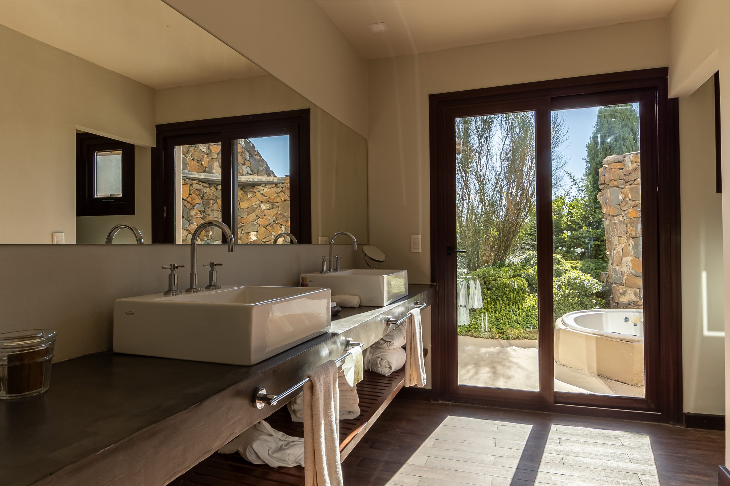 Modern bathroom with double sinks, large mirror, wood accents, and glass doors opening to an outdoor soaking tub.