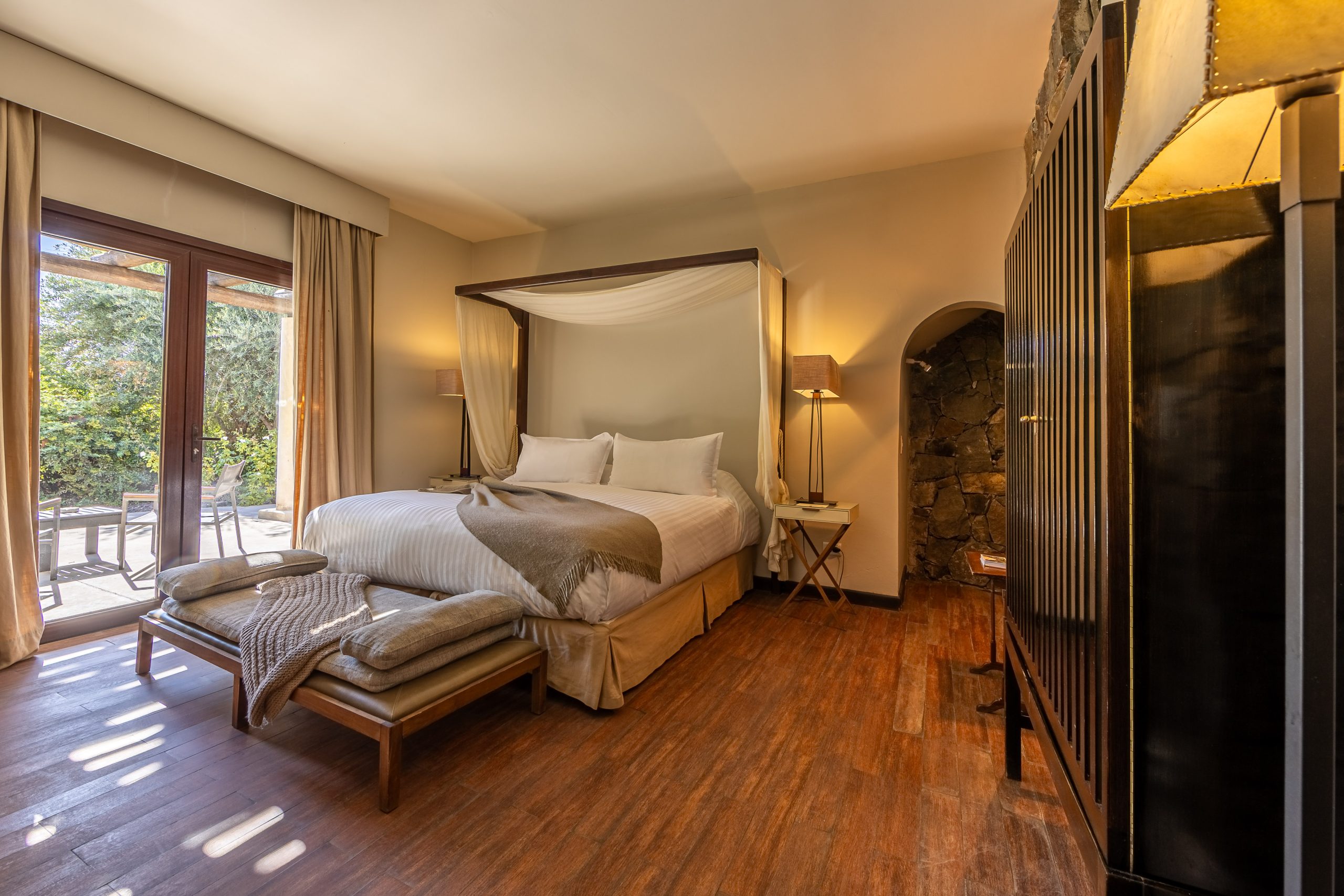 Elegant hotel bedroom with a canopy bed, wooden floors, warm lighting, and a patio view through glass doors.