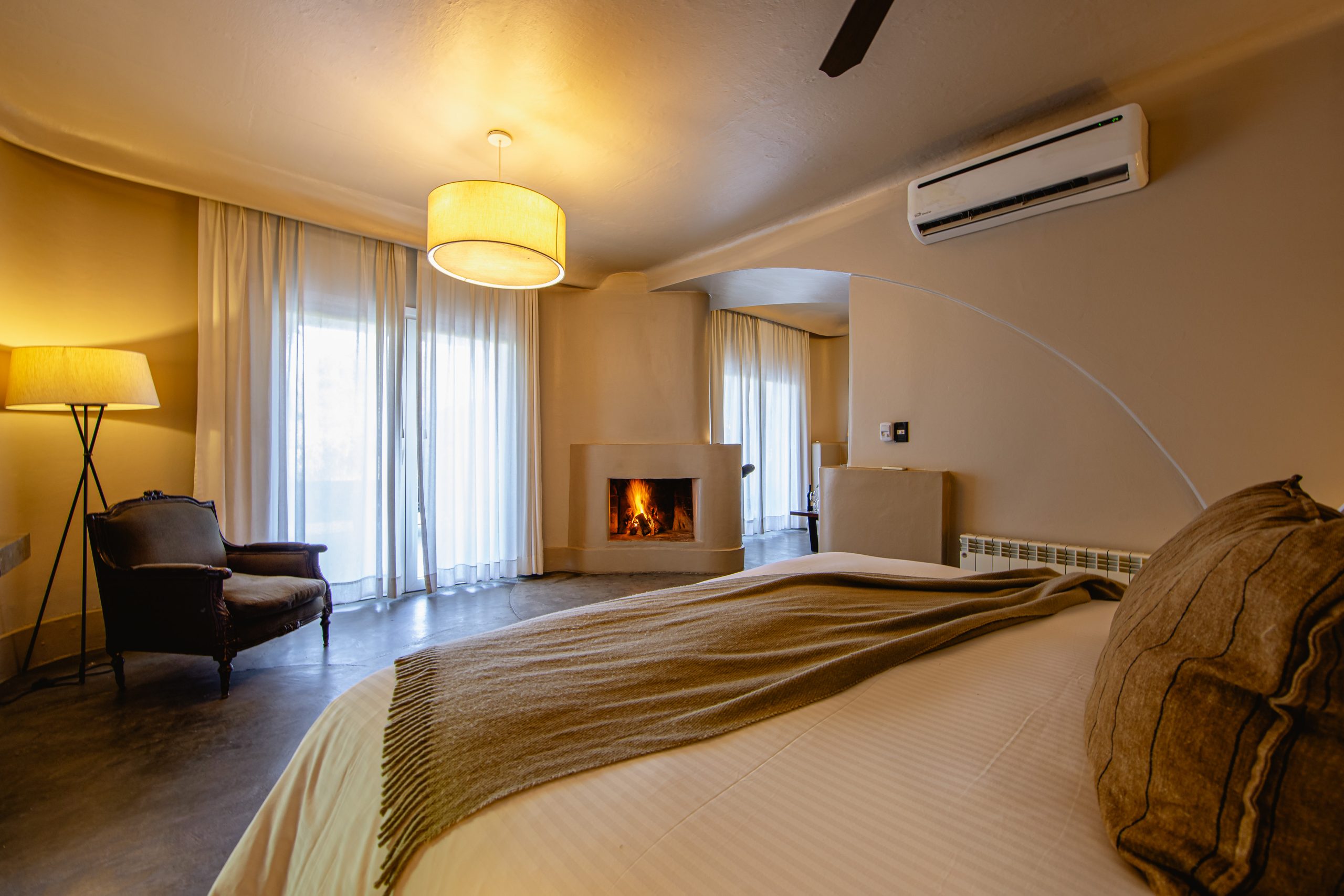 Cozy bedroom with a large bed, warm lighting, fireplace, armchair, and sheer curtains letting in daylight.