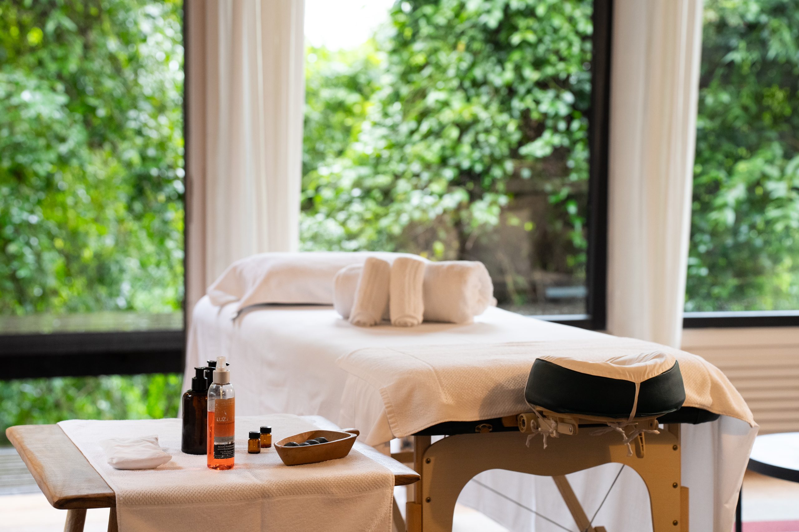 Massage table with folded towels and oils in a serene spa room overlooking lush greenery.