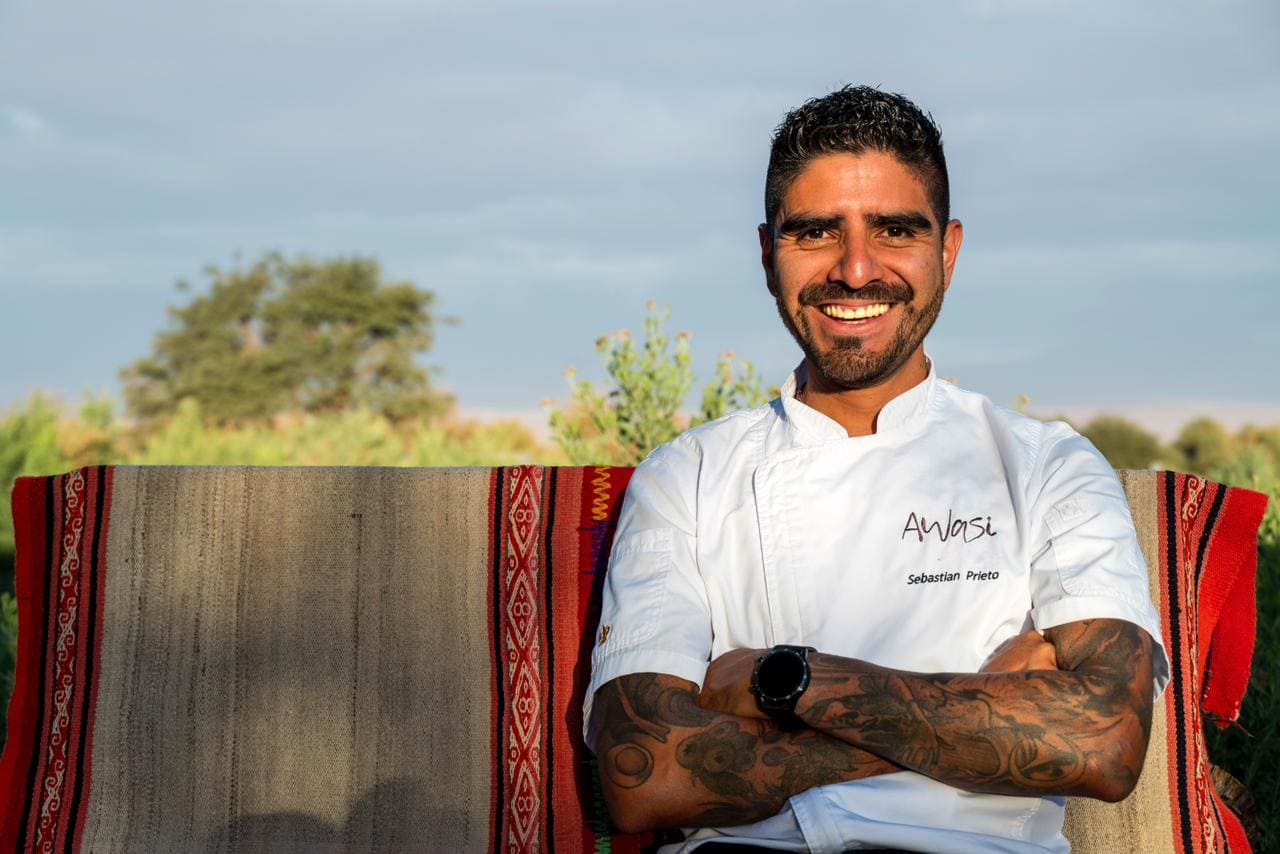 Smiling chef in white Awasi jacket with tattooed arms, seated outdoors against woven textiles and greenery.