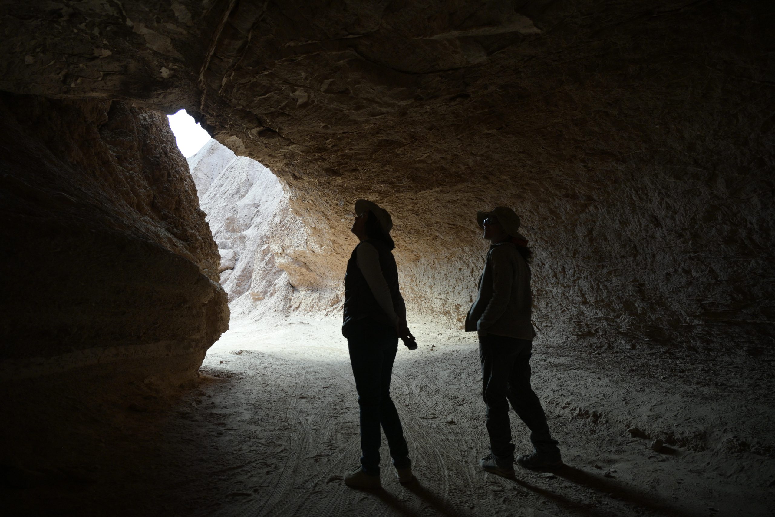 Two people exploring a dark cave, silhouetted against the sunlight streaming in from the entrance, with rugged rock walls surrounding them.