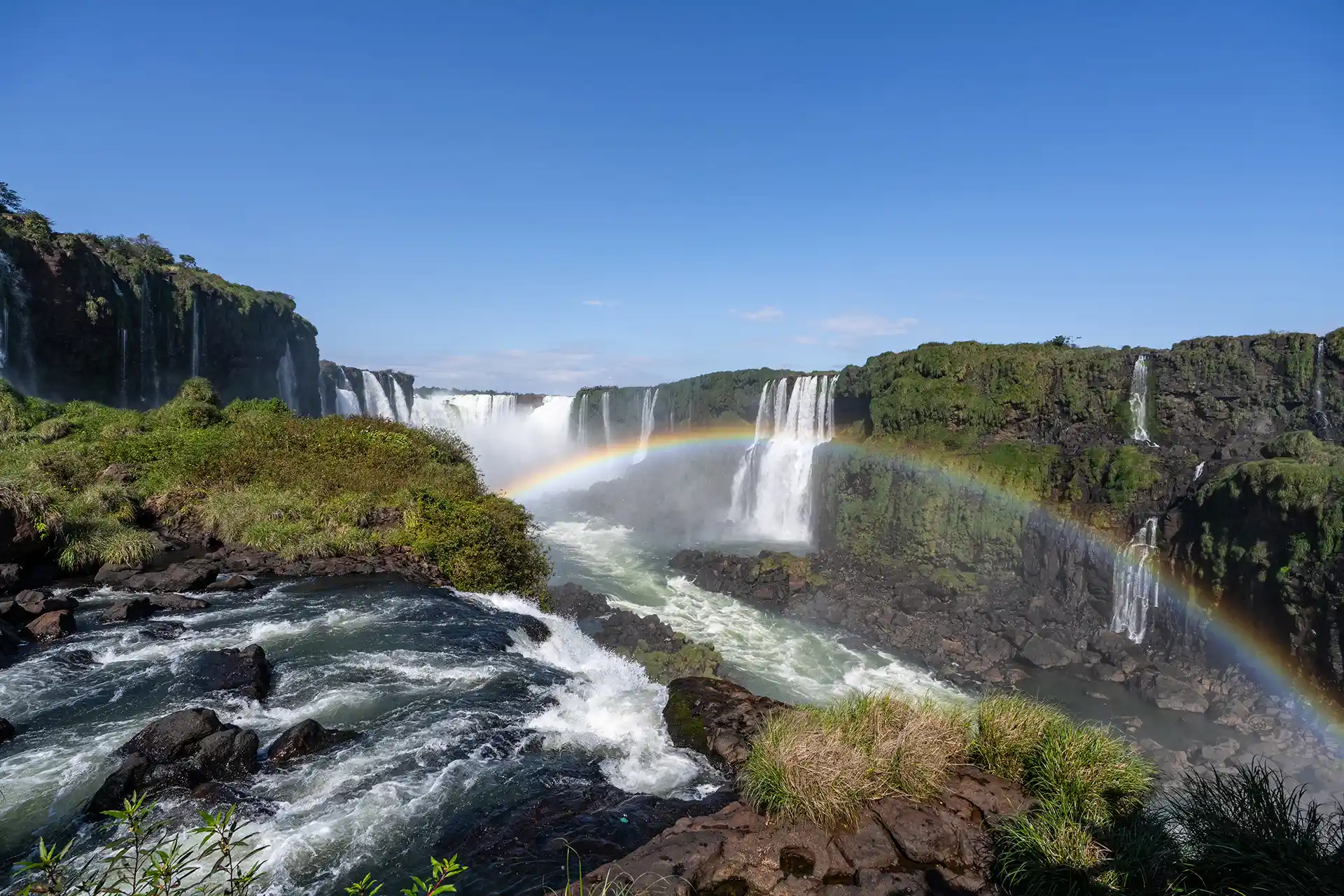 Wide view of cascading waterfalls stretching across lush green cliffs beneath a clear blue sky.