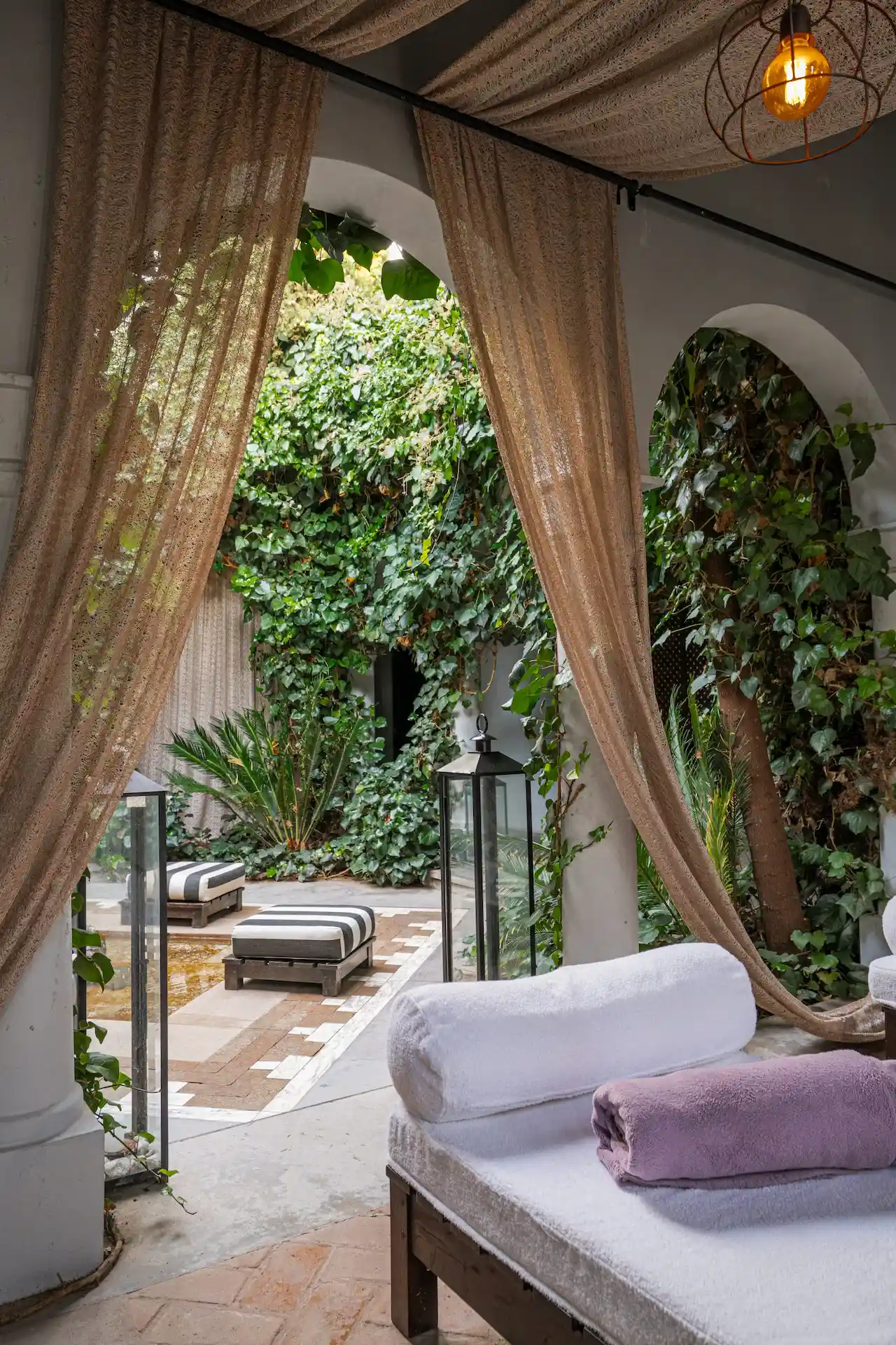 Outdoor spa lounge with cushioned daybeds, sheer curtains, lanterns, and lush greenery in a serene courtyard.