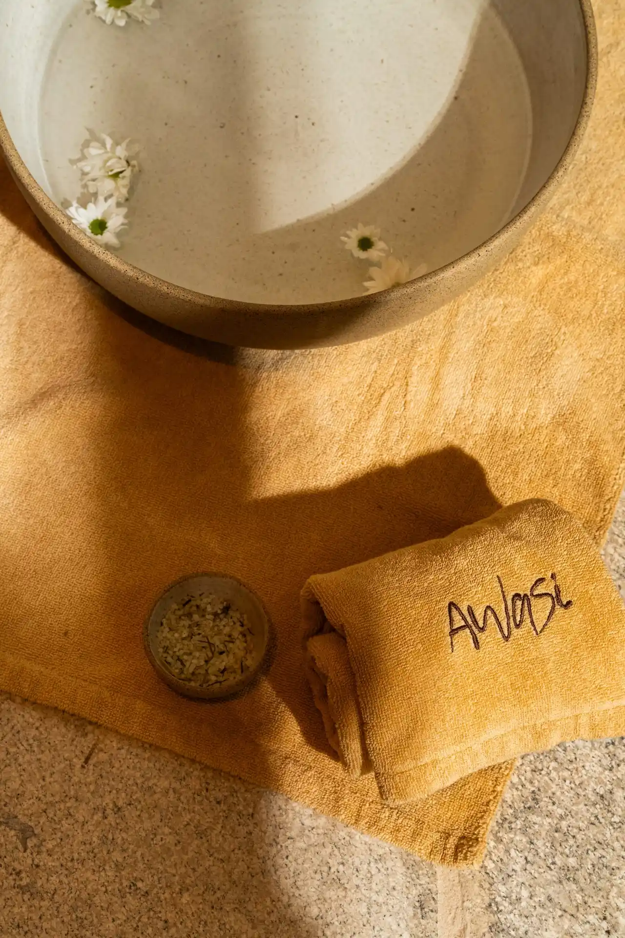 Ceramic bowl with water and floating daisies beside folded anasi towel and bath salts on a warm textured surface.