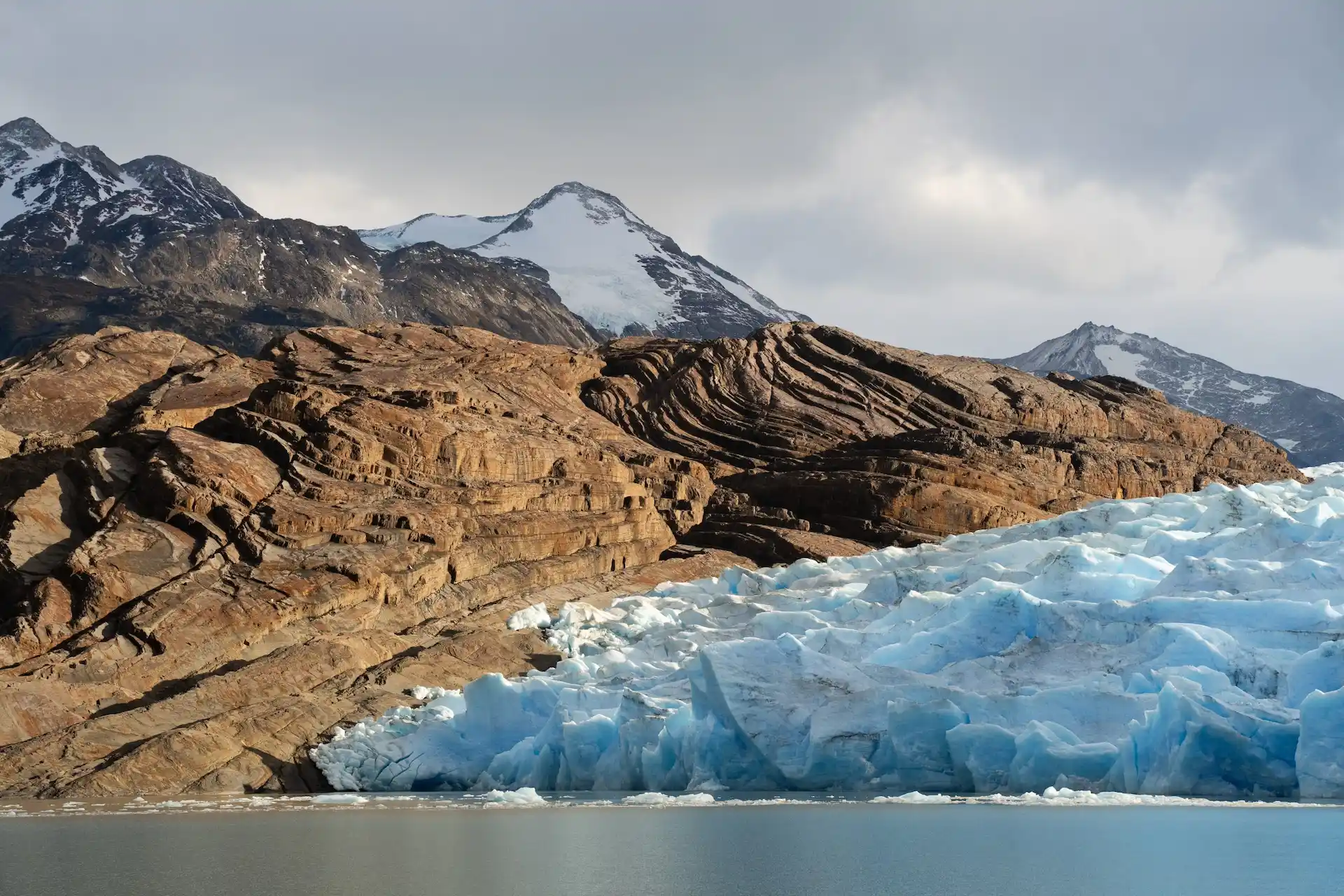 Blue glacier meets rocky mountains and calm water, with snow-capped peaks under a soft, overcast sky.