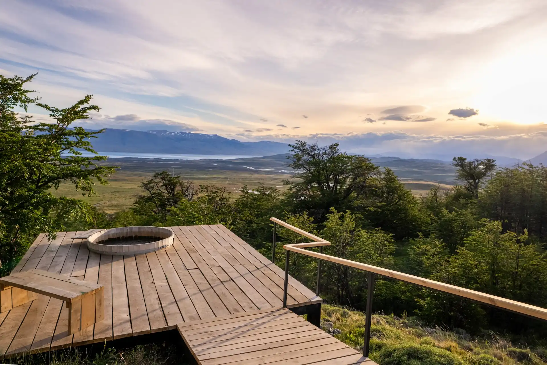 Wooden deck with outdoor tub overlooking vast landscape with mountains forest and lake at sunset
