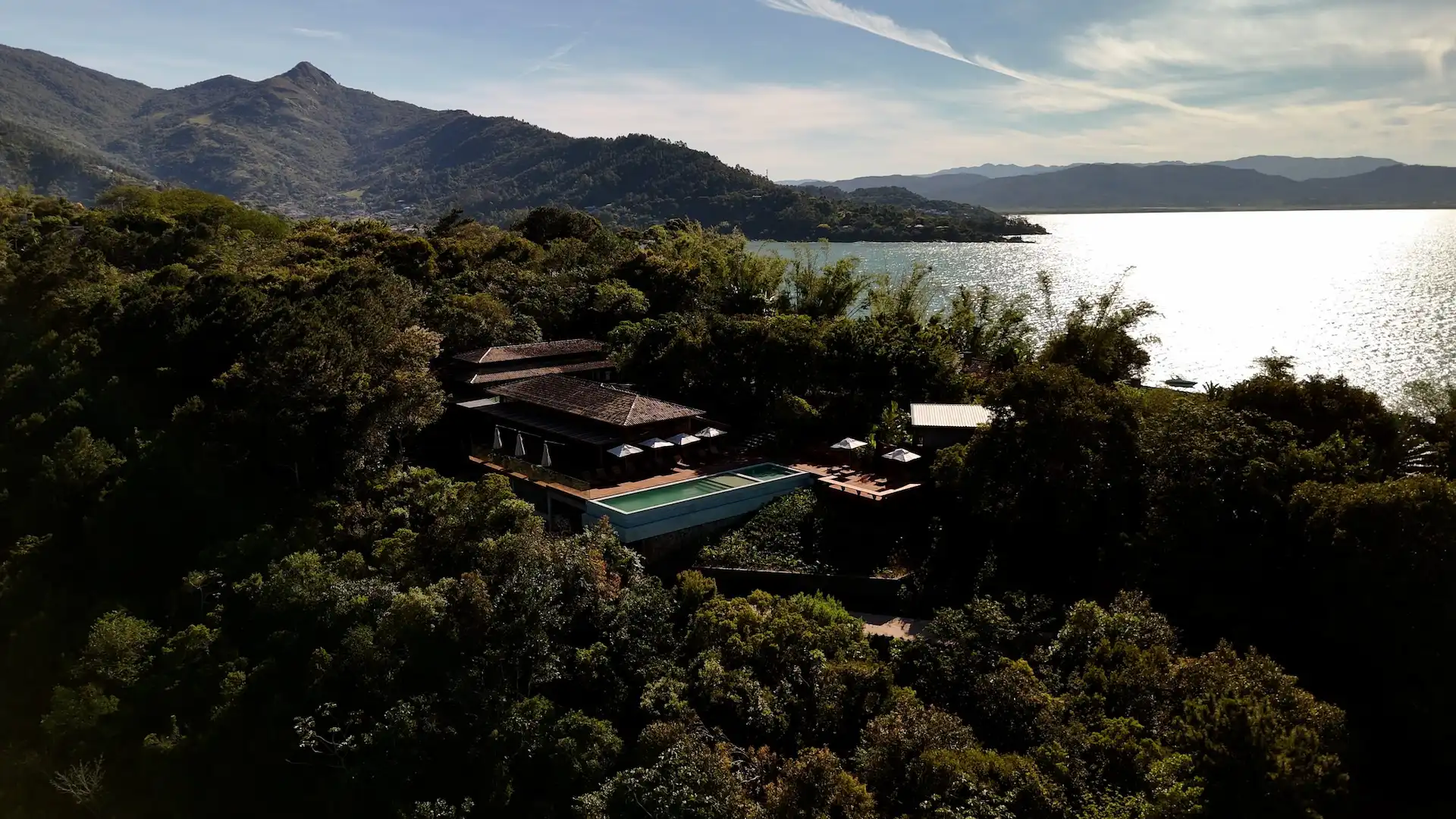 Aerial view of a luxury resort surrounded by forest overlooking a lake with mountains and bright sky