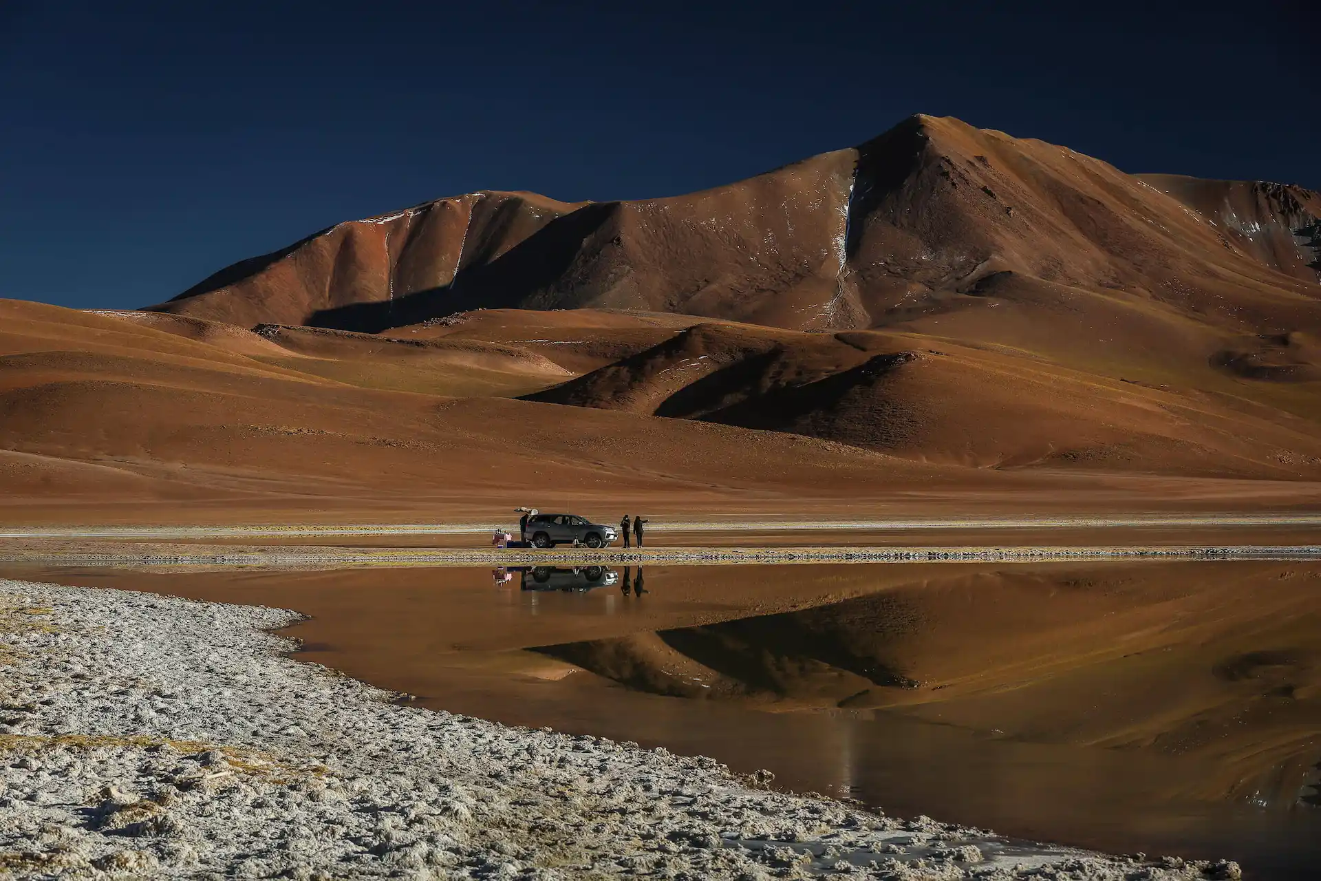 Travelers beside an suv at a reflective desert lake, with rust-colored mountains under a dark sky.